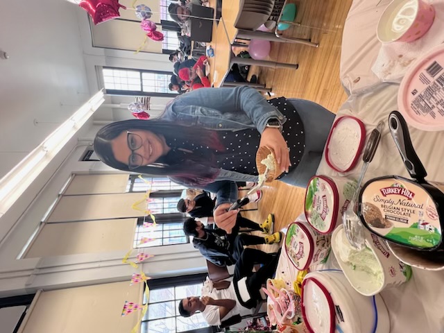 Ms. Araujo posing as she scoops ice cream in back of several open Turkey Hill ice cream containers, and students in distant background behind her