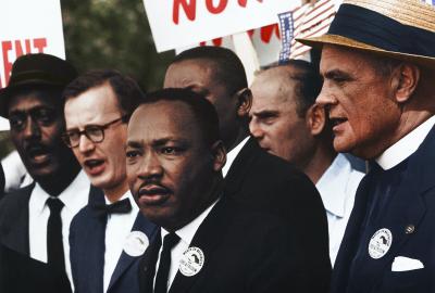 Dr. Martin Luther King Jr. giving a speech at a civil rights protest accompanied by 4 other men and protestors with signs