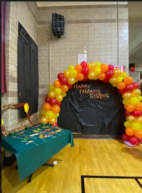 A Photobooth set  up in the gym: Yellow, orange, and red balloon arch over black background with the words "Happy Thanksgiving" 