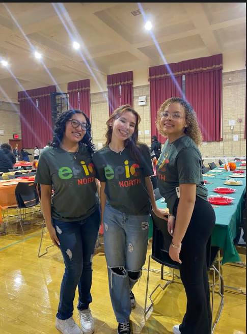 3 female students pose with green EPIC North t-shirts in front of potluck tables in the gymnasium
