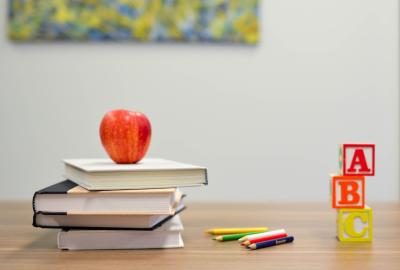 Teachers desk with a red apple on top of 4 stacked books, next to an assortment of colored pencils